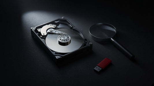 Opened hard drive showing its platter beside a magnifying glass and a red USB stick on a dark desk